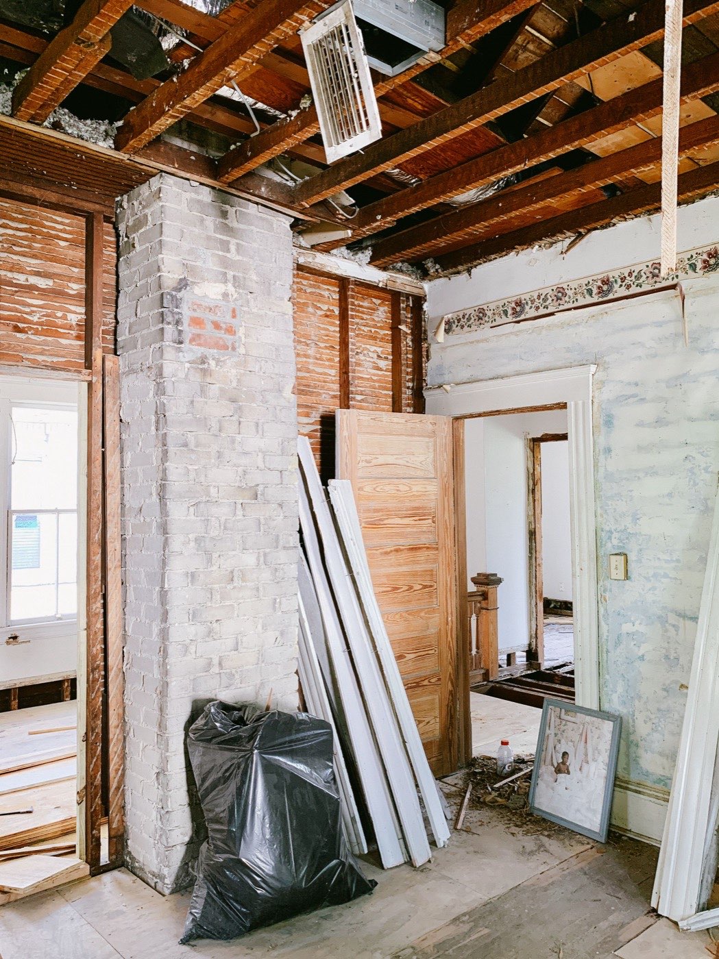 Room mid-demolition with exposed studs and lath, a whitewashed brick chimney, and salvaged doors set aside