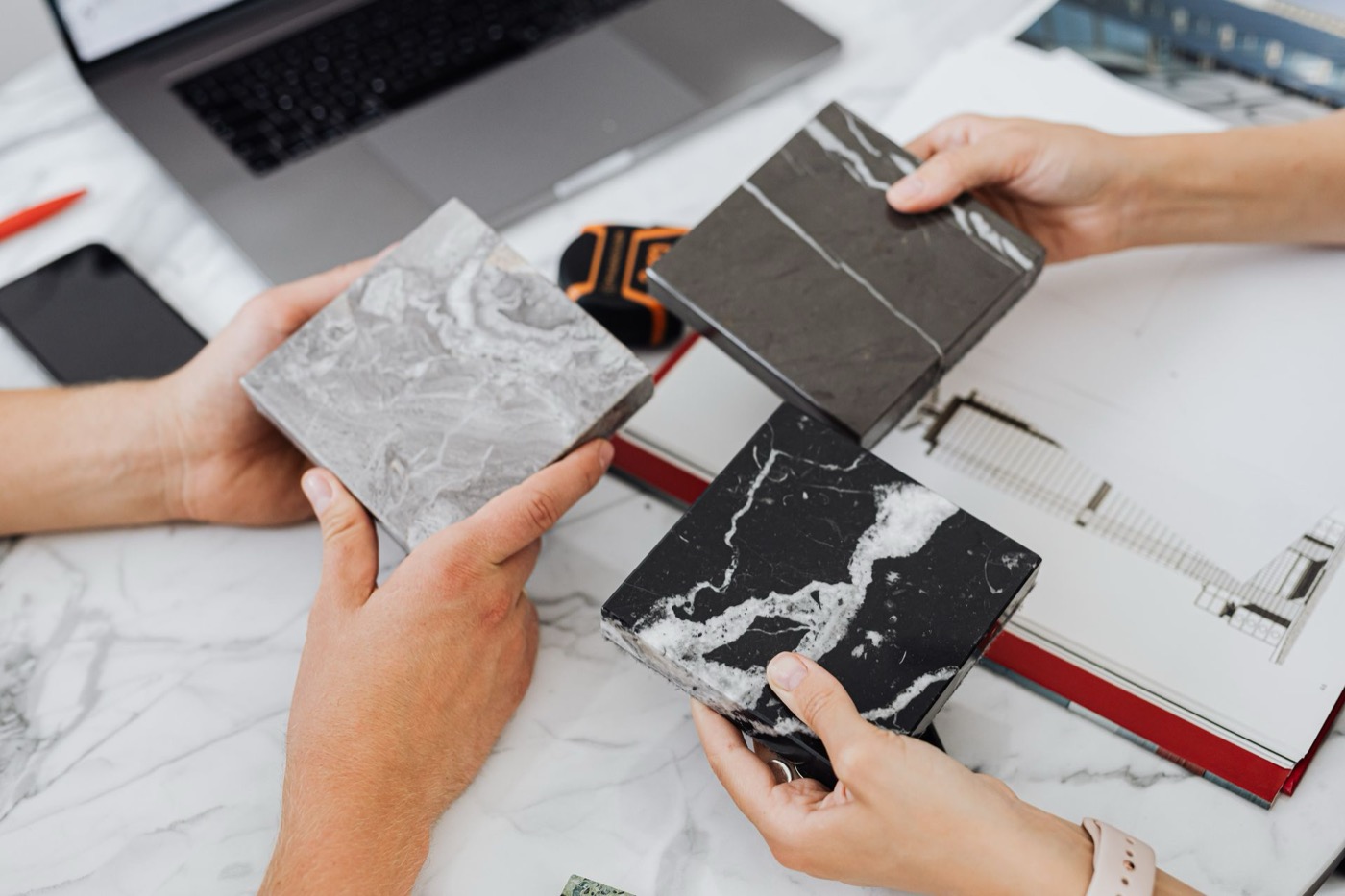 Two people comparing marble tile samples over a desk with blueprints and a laptop